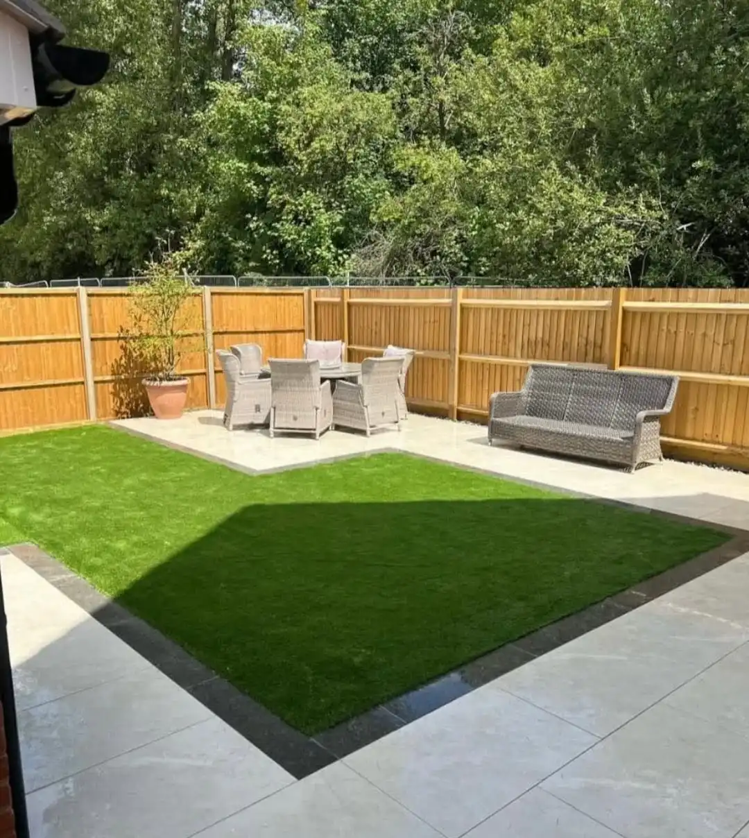 Backyard patio with a central green lawn bordered by dark trim and surrounded by light stone tiles, featuring a wooden fence background, a gray outdoor sofa on the right, and a round patio table with four gray wicker chairs and cushions on the left near a potted plant.