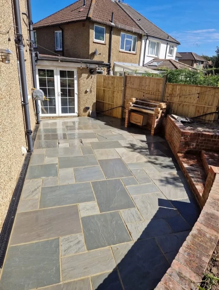 Newly paved backyard patio with large rectangular and square stone tiles in varying shades of gray and beige, adjacent to a brick barbecue grill and enclosed by a wooden fence, with a house featuring beige roughcast walls and white-framed glass doors.