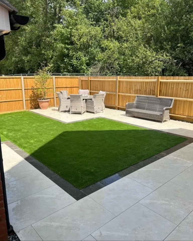 Backyard with a square grass lawn bordered by dark trim, surrounded by light-colored tiled patio, wooden fencing, outdoor dining table with four chairs, a bench, and a potted plant, all under sunny daylight with trees in the background.