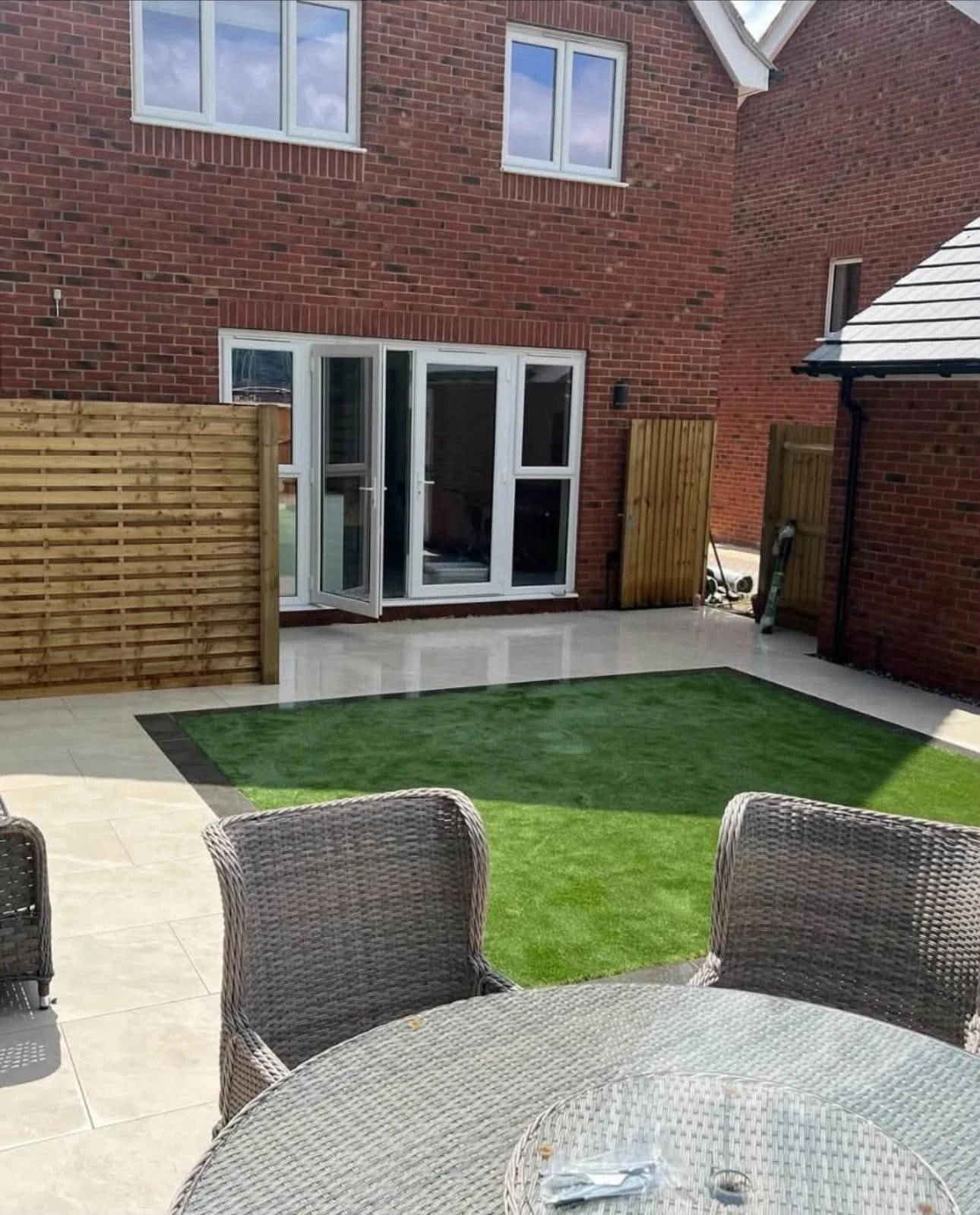 Backyard patio with beige tiles, a central square of artificial grass bordered by dark bricks, surrounded by a wooden fence and adjacent to a red brick house with open white-framed double doors and windows, and gray wicker chairs around a round glass-top table.