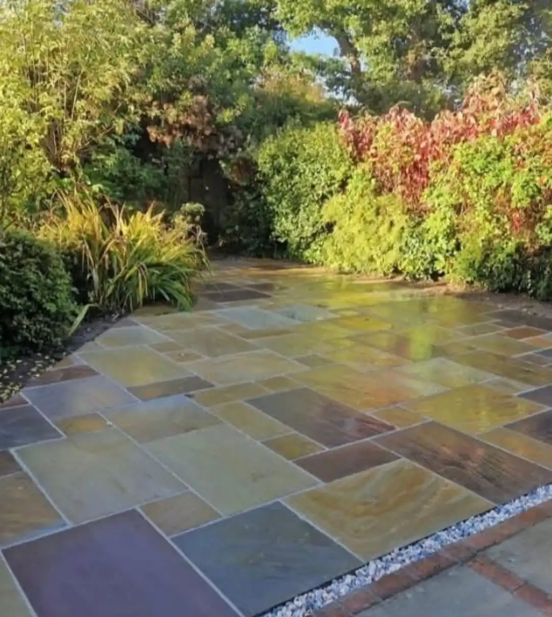 A sunlit stone patio with multi-colored rectangular slabs bordered by bricks and white gravel, surrounded by lush green and red foliage.