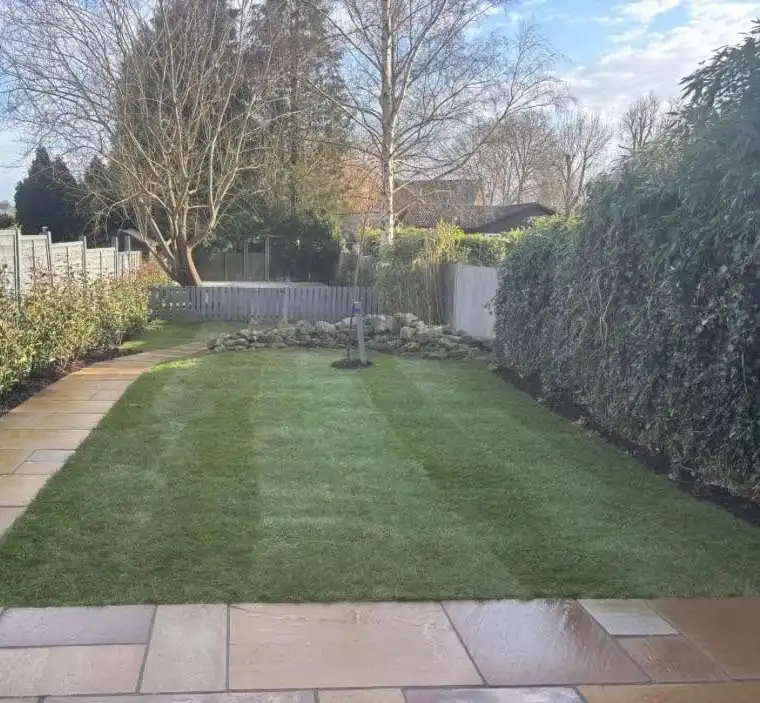 A neatly mowed rectangular lawn bordered by a tiled walkway on the left and trimmed shrubs on the right, with a pile of rocks and leafless trees in the background under a partly cloudy sky.