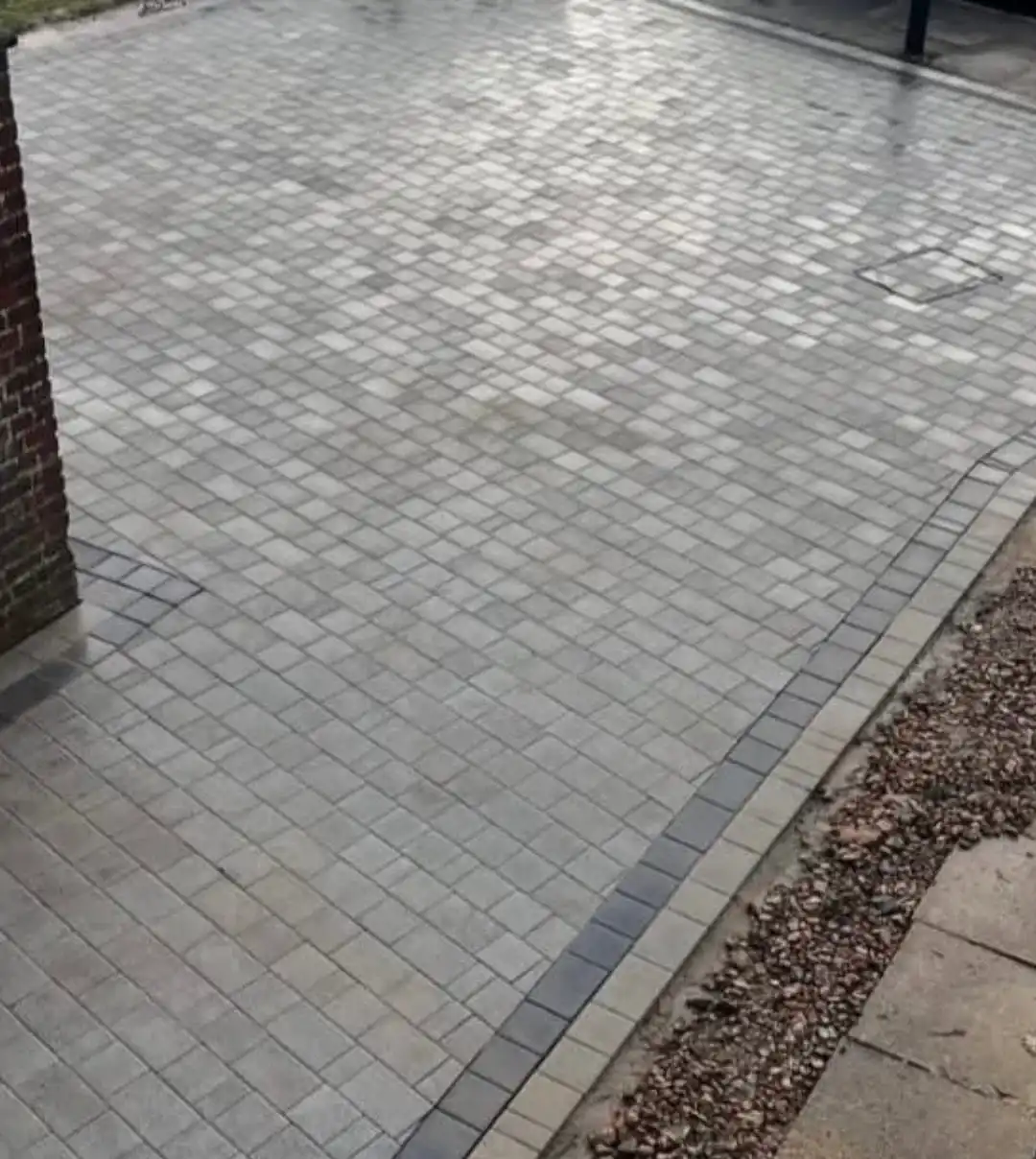 Gray paved driveway with rectangular and square stones in a pattern, bordered by darker stones on one side and a gravel strip on the other.