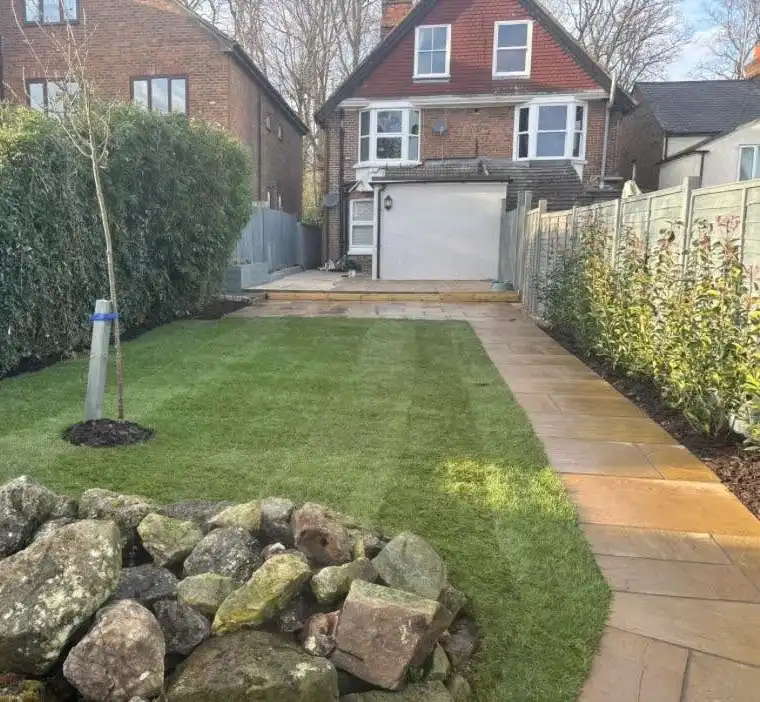 Backyard with neatly trimmed grass, a small young tree supported by a stake, a rock arrangement in the foreground, stone pathway along the right side, tall hedges on the left, and a brick house with a closed garage door and windows in the background.