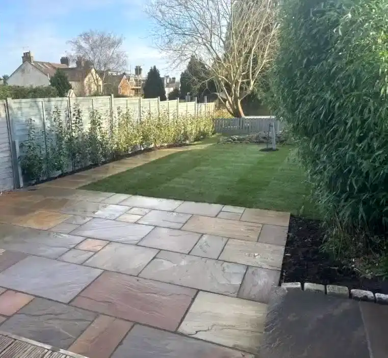 A backyard with a paved stone patio in the foreground, bordered by a green lawn and a row of small trees along a grey fence, with leafless trees and houses in the background under a clear sky.