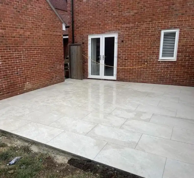 Outdoor patio area with newly installed large light beige rectangular tiles and red brick walls with a white-framed glass door and a window with shutters.