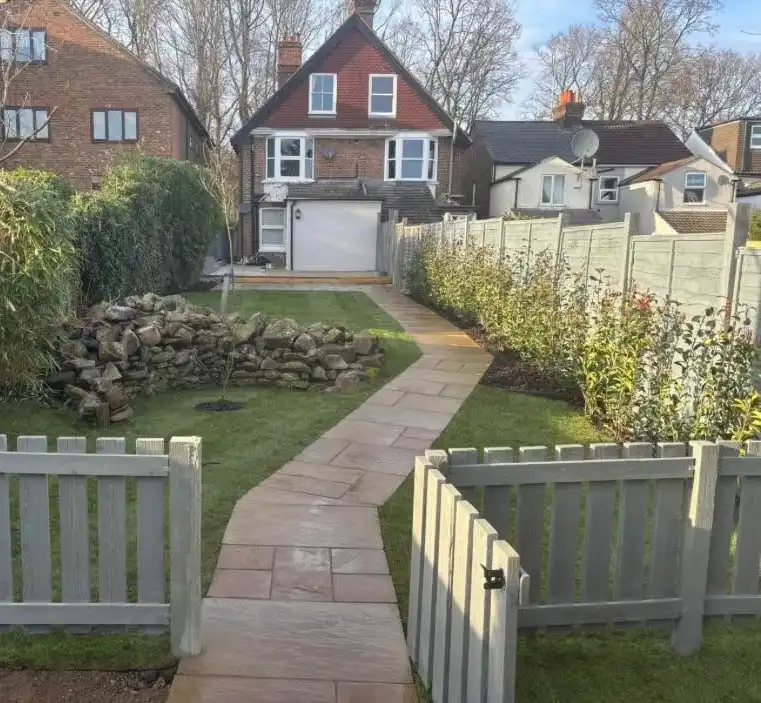 Backyard garden with a curved stone pathway leading to a two-story brick house with white trim and a garage door, flanked by green hedges, a stone pile, and a wooden fence with an open gate in the foreground.